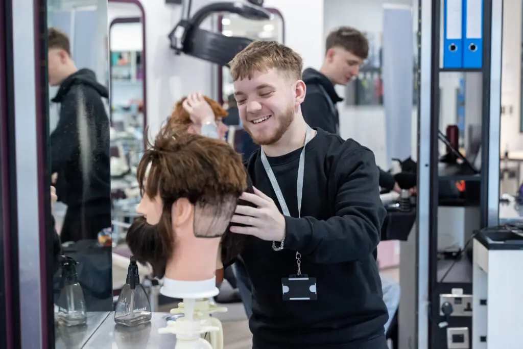 A student smiles while practising a haircut on a mannequin head in a college hairdressing salon.