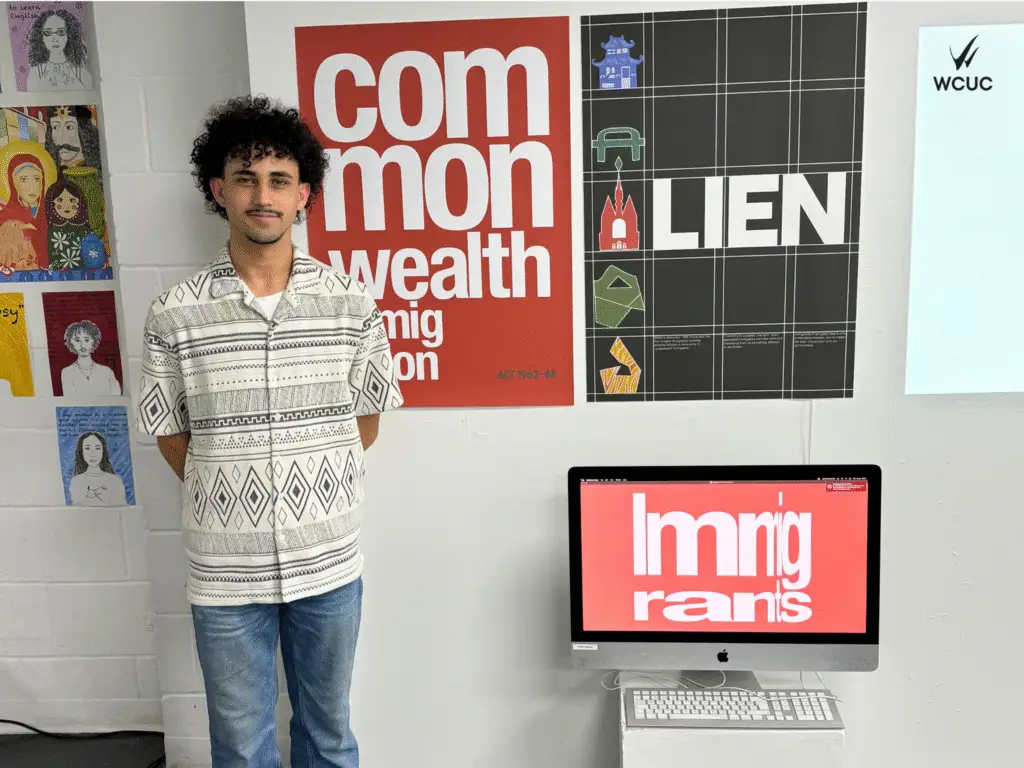 A student stand in front of his graphic design project exploring immigration, featuring bold red and black posters and a desktop screen with the word "immigrants" in distorted text. 