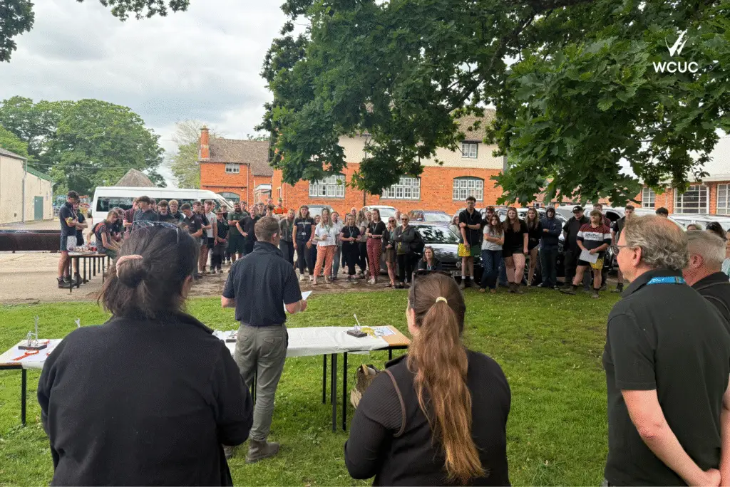A tutor speaks to a large gathered audience of students and staff at the Lackham awards ceremony, standing beside the rosette and trophy table.