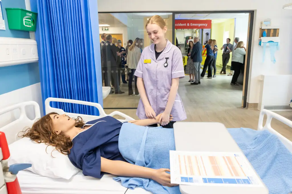 A student nurse practices bedside care with a medical mannequin in a simulated hospital ward, while visitors observe through a glass window. 