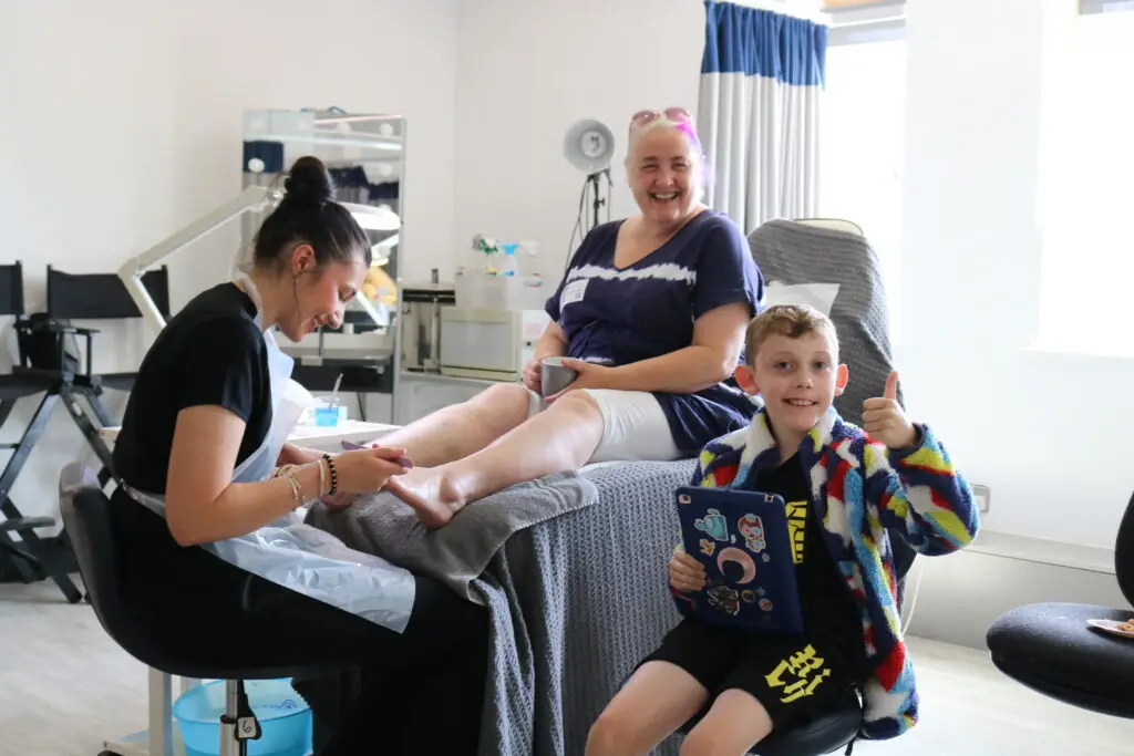 A student gives a pedicure to a smiling woman seated on a treatment couch, while a cheerful young boy beside her gives a thumbs up and holds a tablet decorated with stickers.