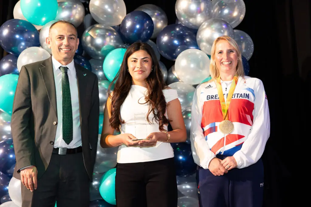 A student in a white top holds an award between a suited man and a Team GB Paralympian wearing a gold medal, all smiling in front of a colourful balloon wall.