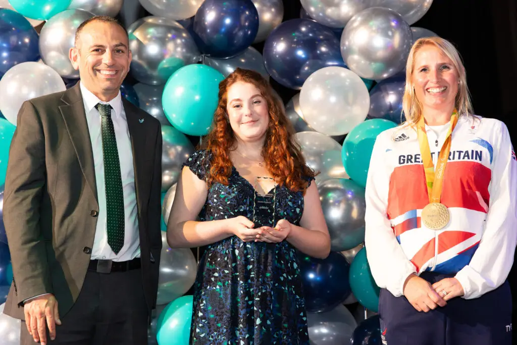 A student holding an award stands between a suited man and a smiling Team GB Paralympian in front of a decorative balloon backdrop.