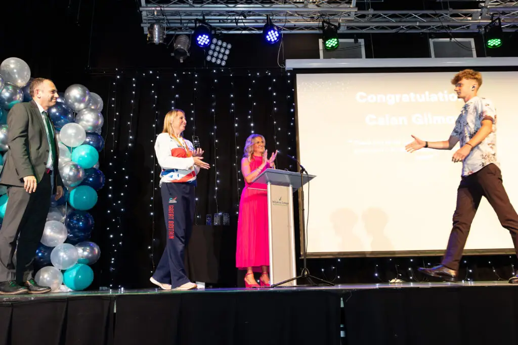 A student walks across the stage to receive an award from a Team GB Paralympian, who holds the trophy. The event host and a suited man stand clapping near the podium.