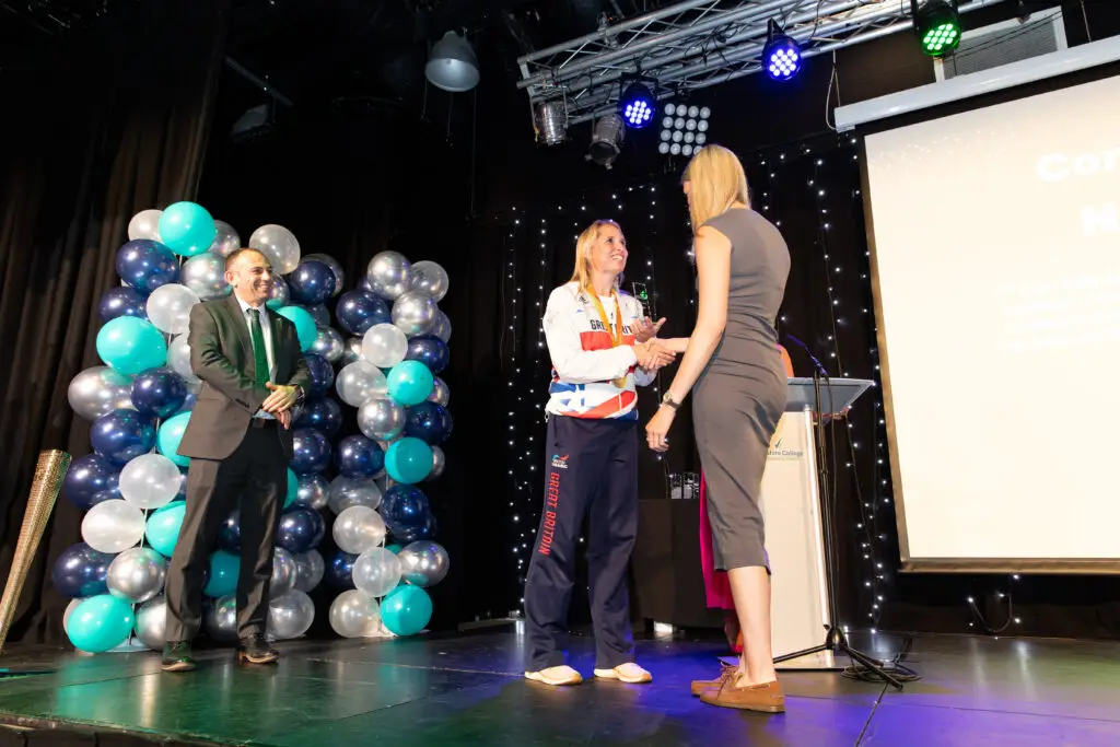 A student receives an award on stage, shaking hands with a smiling Team GB Paralympian. A suited man and the event host applaud from nearby.