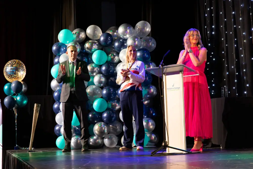 Three presenters on stage — a suited man, a Team GB Paralympian, and a woman in a bright pink dress - stand clapping in front of a balloon backdrop and a podium bearing the Wiltshire College & University Centre logo.