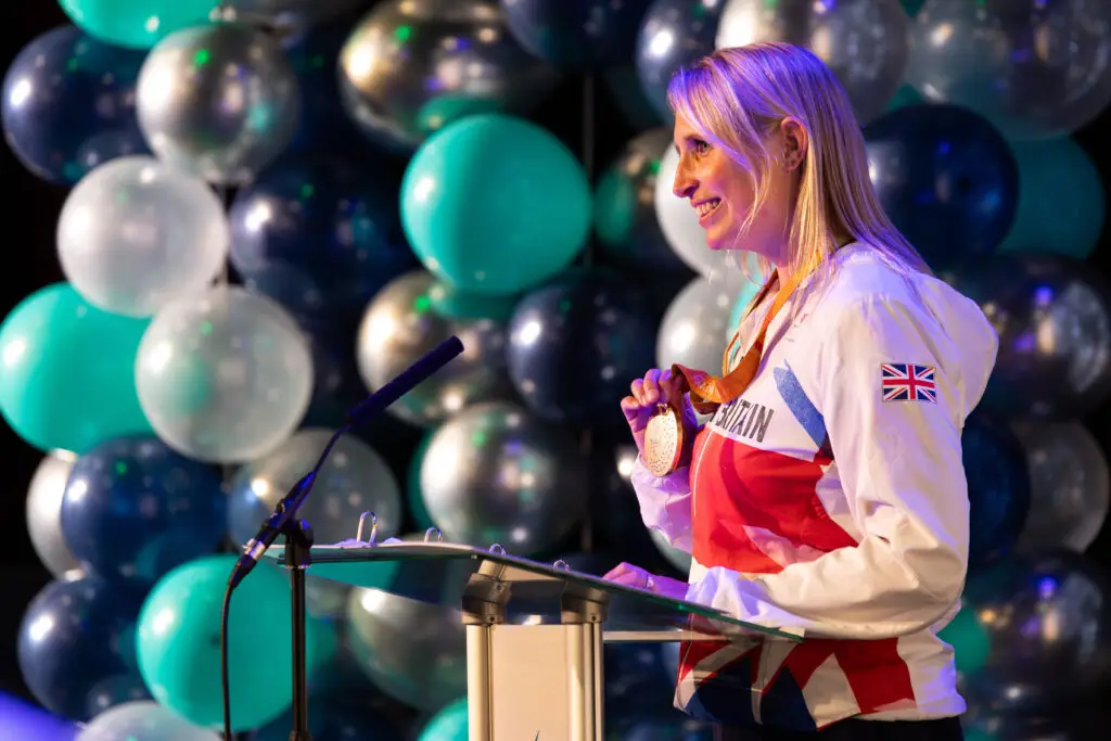 A Team GB Paralympian speaks at a podium while holding up a gold medal, standing in front of a decorative balloon backdrop.