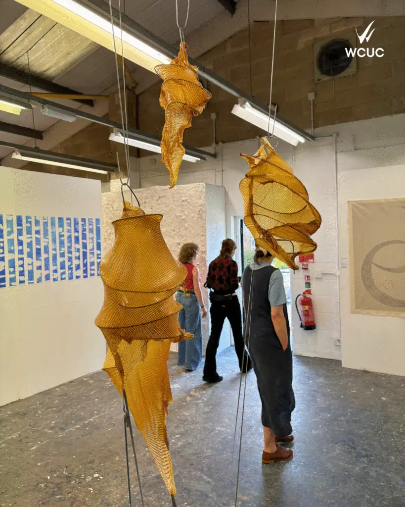 Sculputural forms made from amber mesh fabric hang from the ceiling in a gallery space, with visitors walking and viewing student artworks in the background. 