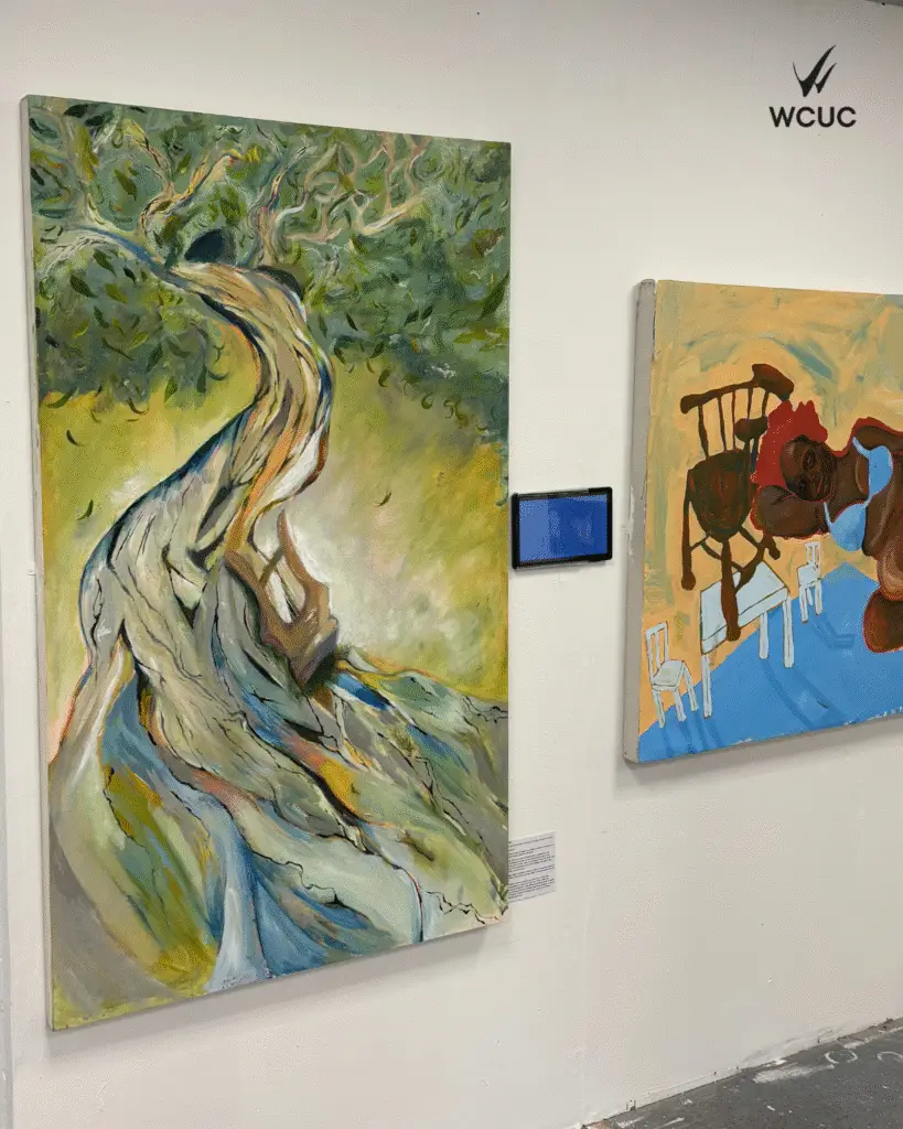 A large oil painting of a twisted, abstract tree with vibrant and yellow tones is displayed on a gallery wall alongside another expressive painting of a seated figure in warm colours. 