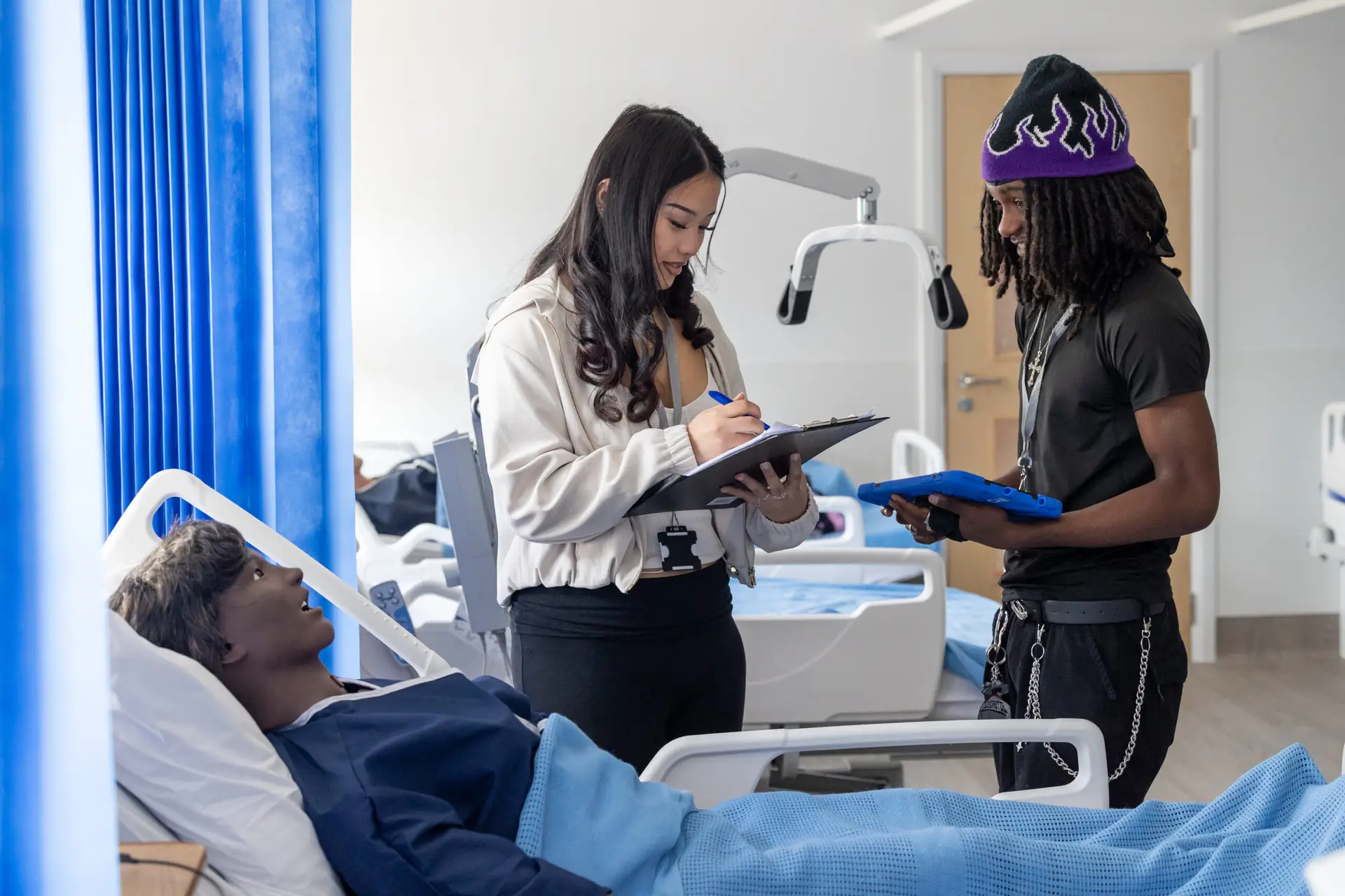 Two students practise recording patient observations using a mannequin in a simulated hospital ward.