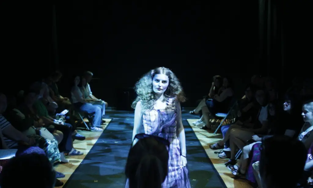 A single model walks down a runway in a long tartan dress with netted purple accents, under blue stage lighting. Her hair is styled in voluminous curls, and she walks towards the camera while the audience looks on.