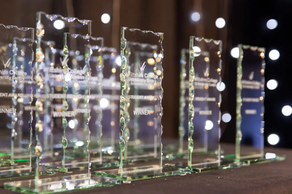 A table laid with a black table cloth holding numerous glass, rectangular trophies in a neat line. The trophies have 'Wiltshire College & University Centre Student Award WINNER' engraved on them. White fairy lights twinkle in the background.