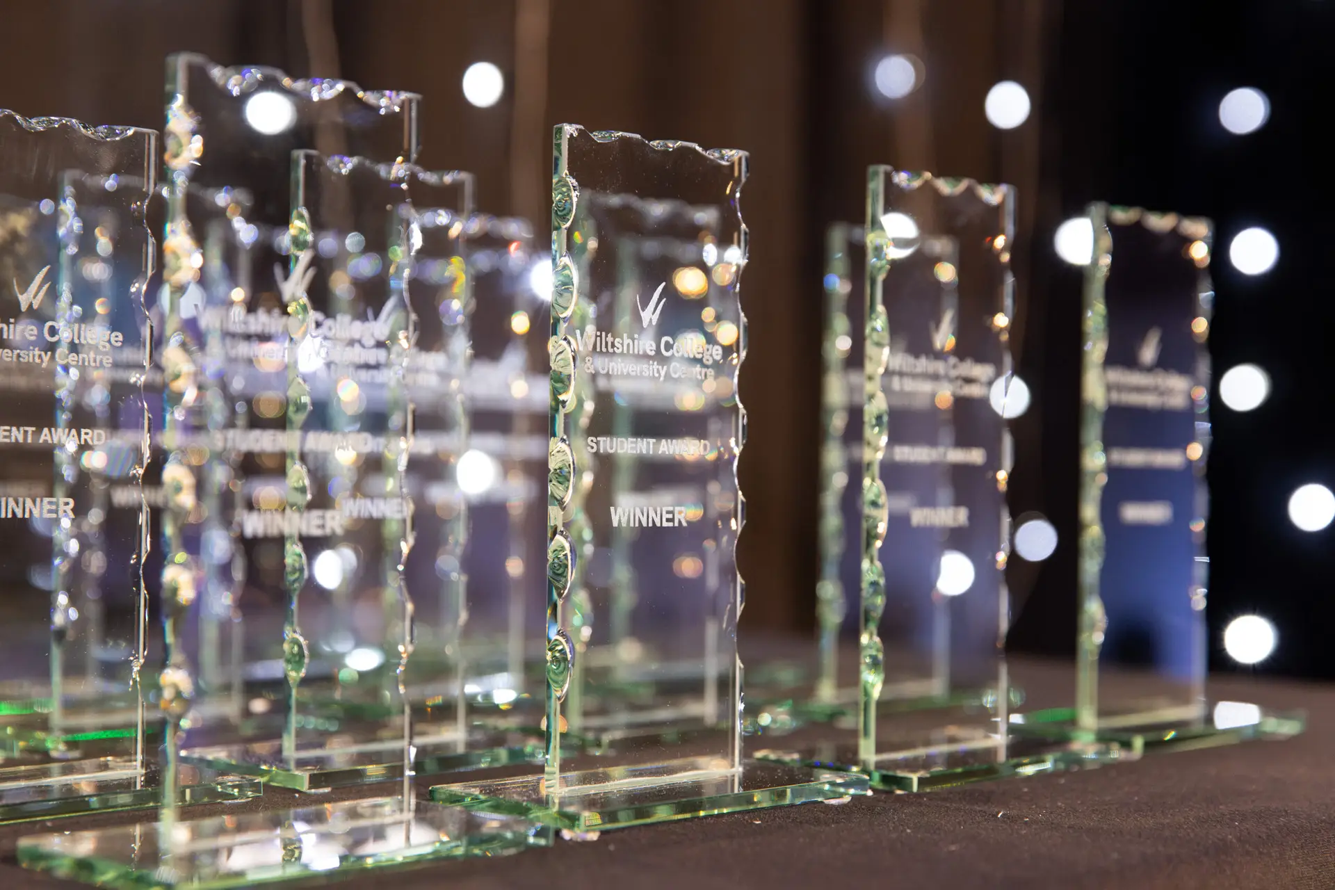 A table laid with a black table cloth holding numerous glass, rectangular trophies in a neat line. The trophies have 'Wiltshire College & University Centre Student Award WINNER' engraved on them. White fairy lights twinkle in the background.