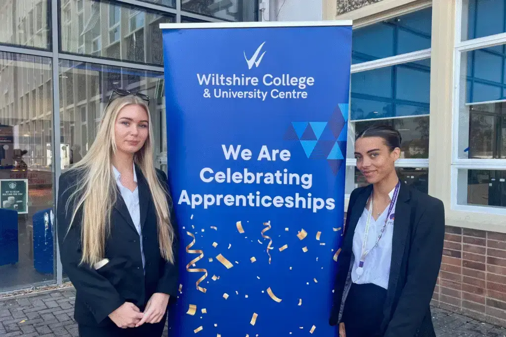 Two young women in business attire standing on either side of a banner reading "We are celebrating apprenticeships" at Wiltshire College & University Centre.