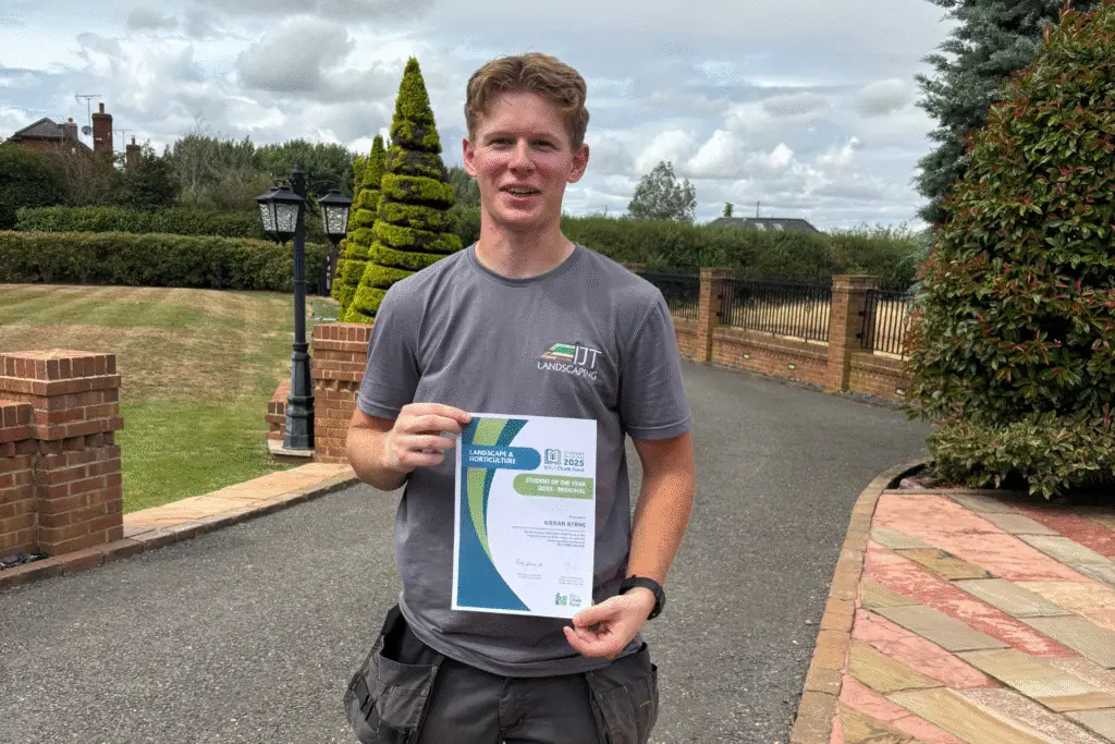 Kieran Byrne, a young man wearing a grey IJT Landscaping t-shirt, stands on a landscaped driveway holding his BALI Chalk Fund Student of the Year 2025 certificate.