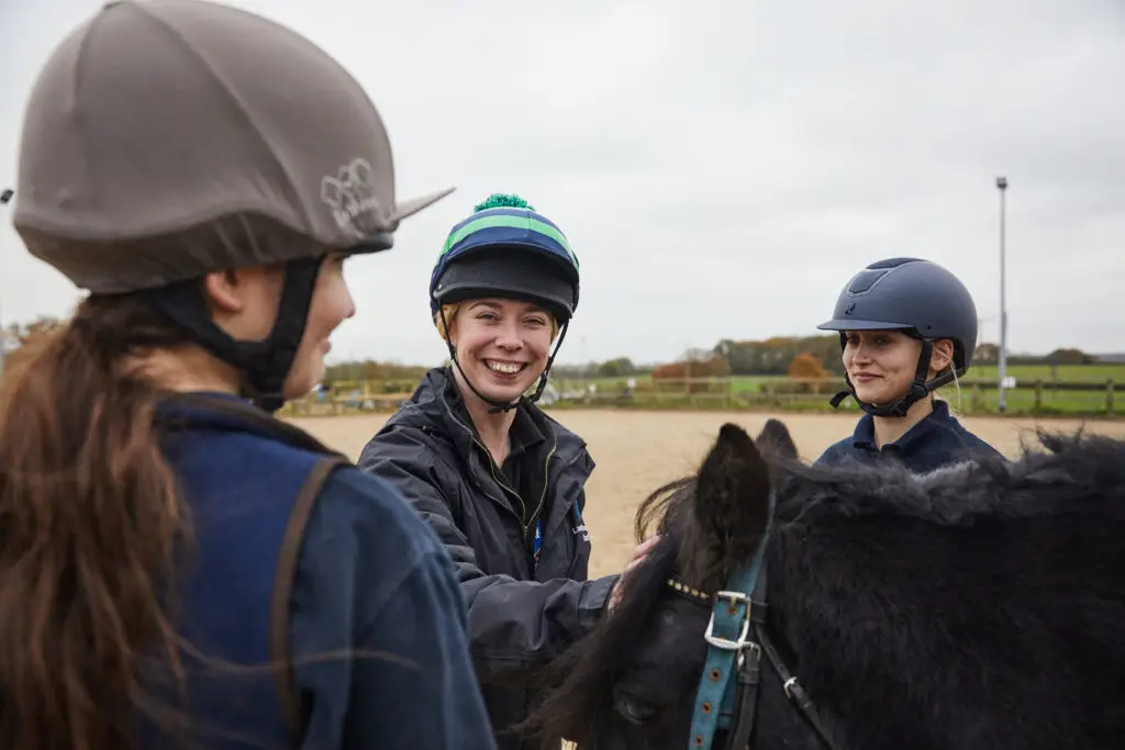 Equine lecturer smiling with two students wearing helmets, standing beside a black horse in an outdoor arena.