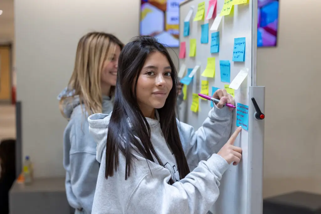 A student in a grey hoodie adds a sticky note to a whiteboard covered in colourful notes, with another student smiling in the background.