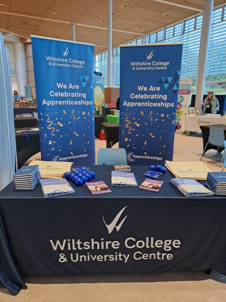 Wiltshire College & University Centre stand with banners reading "We are celebrating apprenticeships" and a display of brochures, tote bags and promotional items.