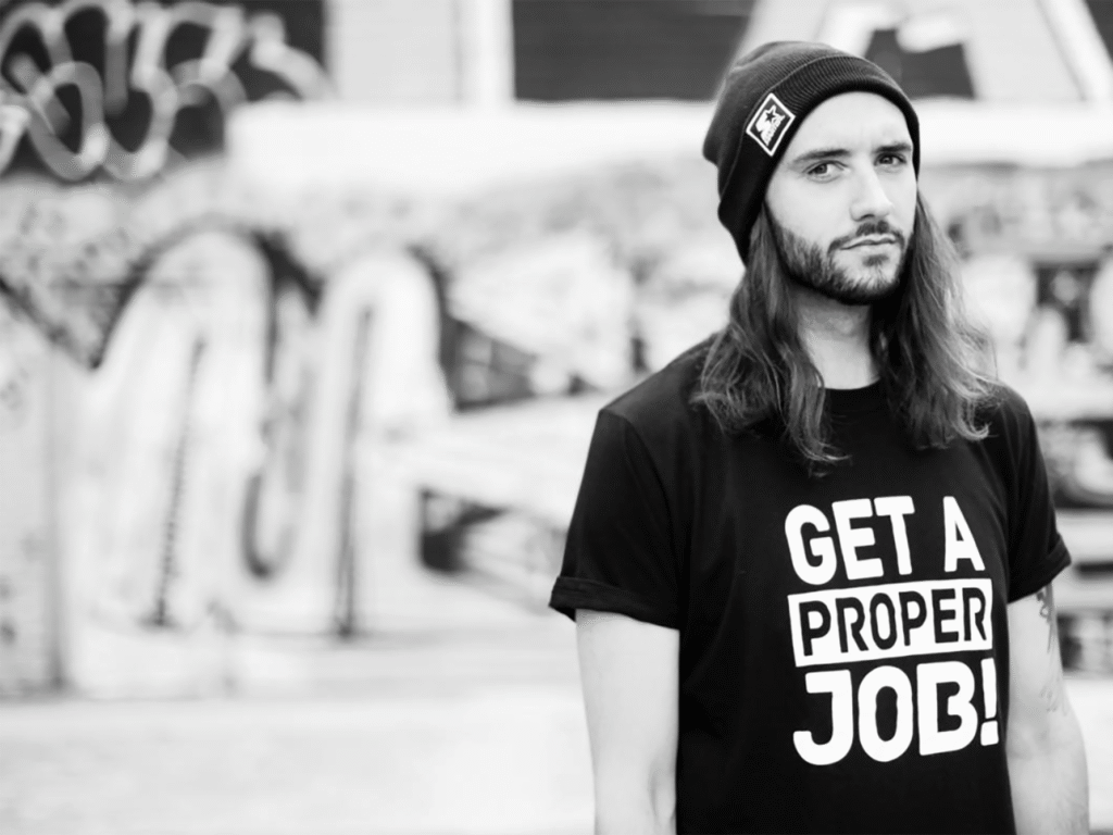 Person wearing a beanie and a black t-shirt with the words "Get a proper job!" standing in front of graffiti.