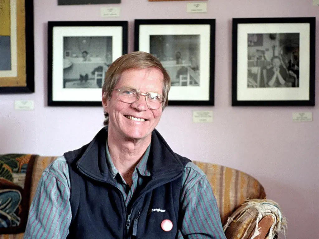 Smiling person with glasses, sitting indoors in front of framed photographs.