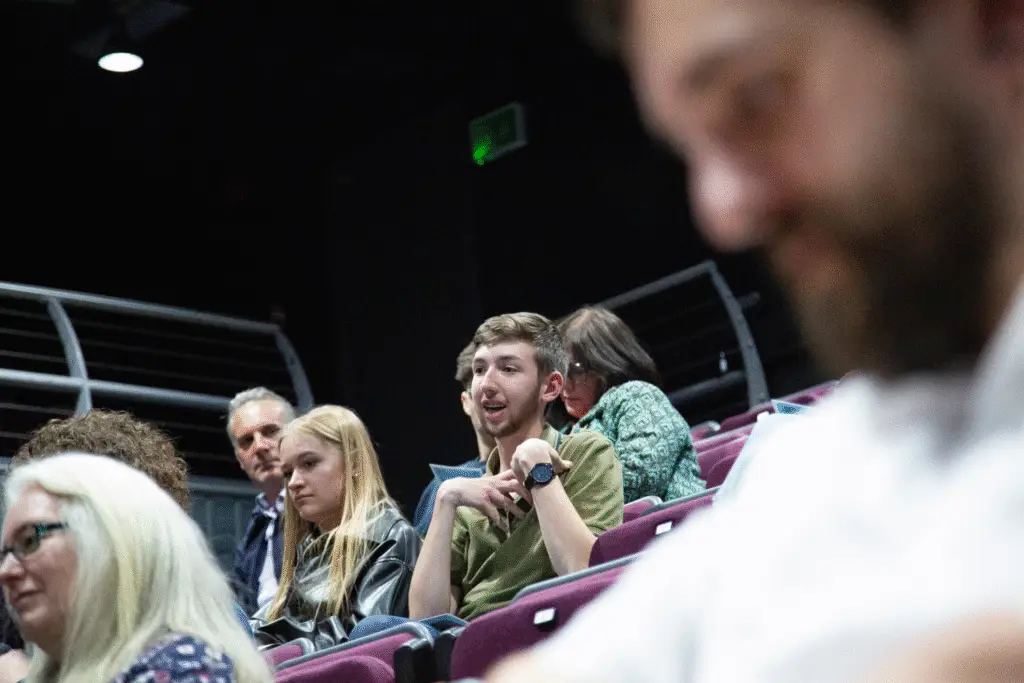 A student speaks while seated among peers in the theatre.