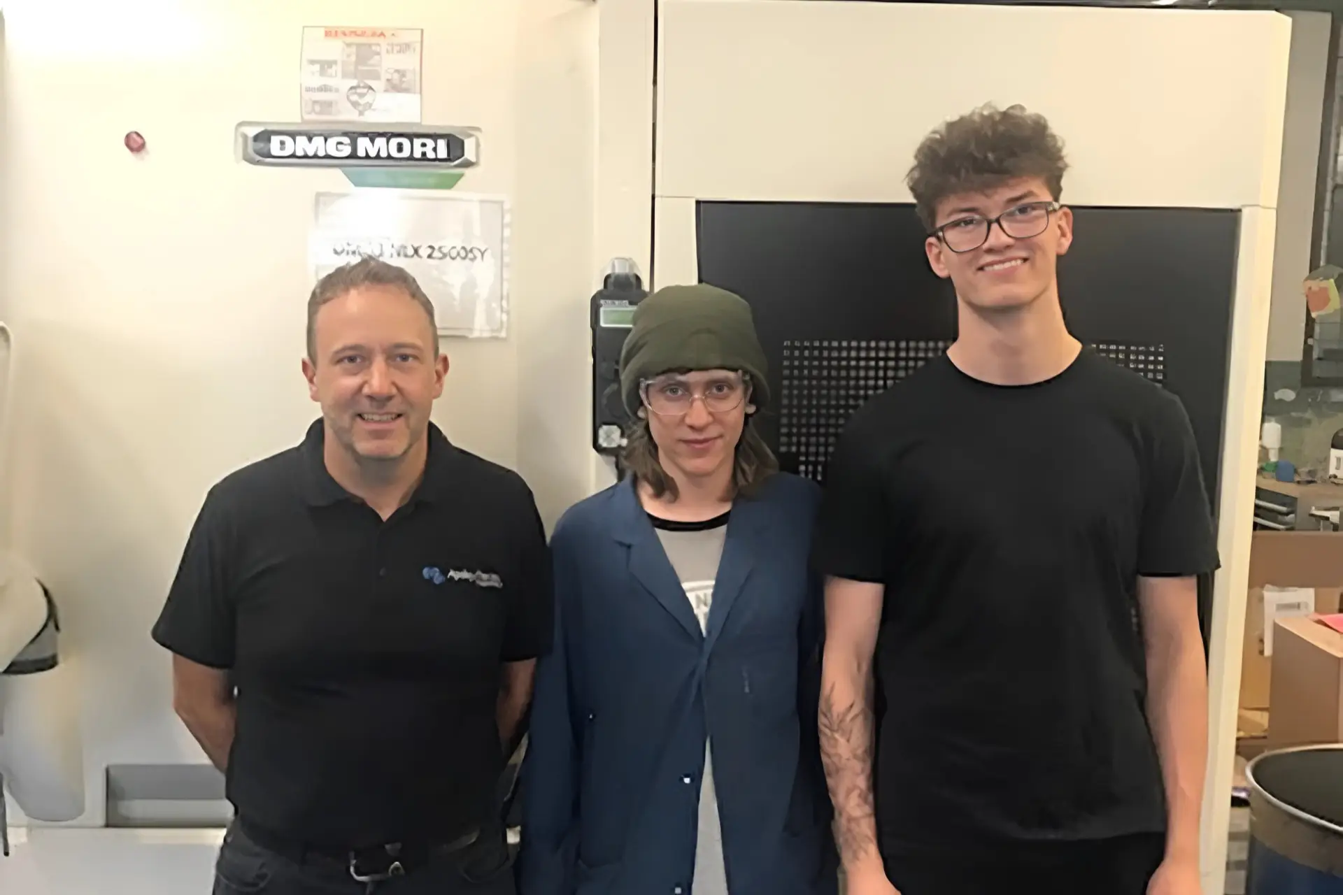 Three people standing in front of engineering machinery at Apsley Precision Engineering, including a staff member and two students on T Level placements.
