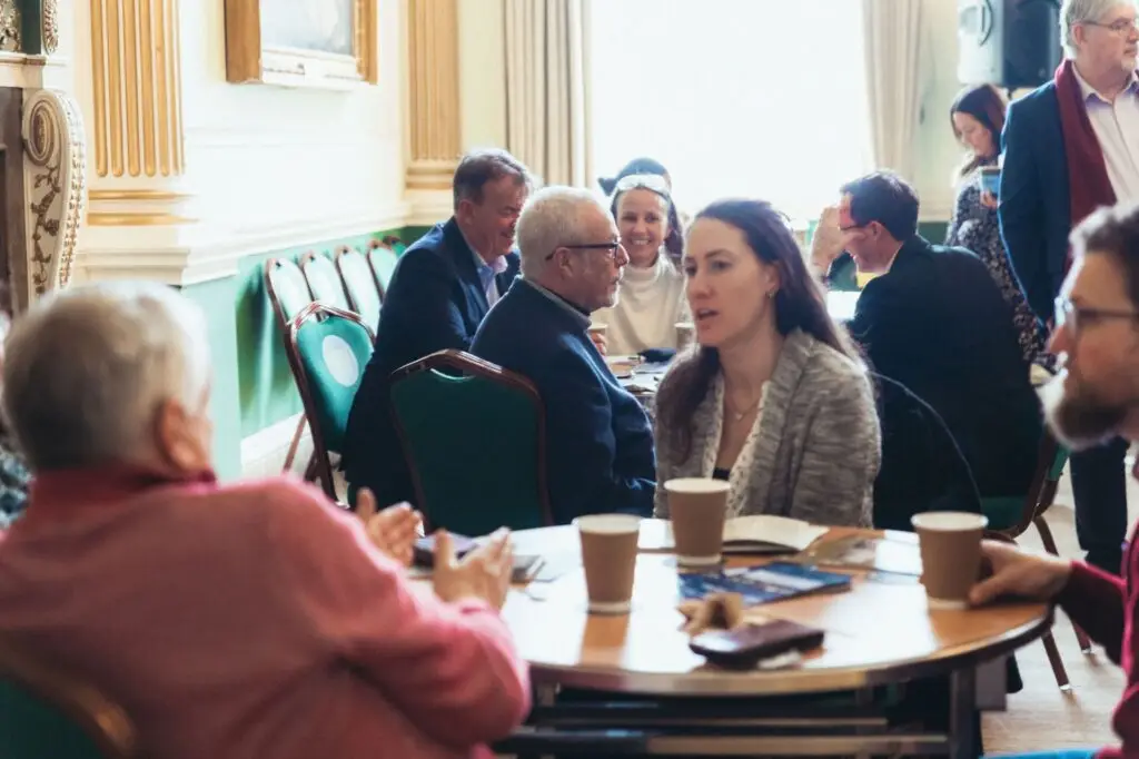 People seated around tables in a bright conference room, talking and networking over coffee.