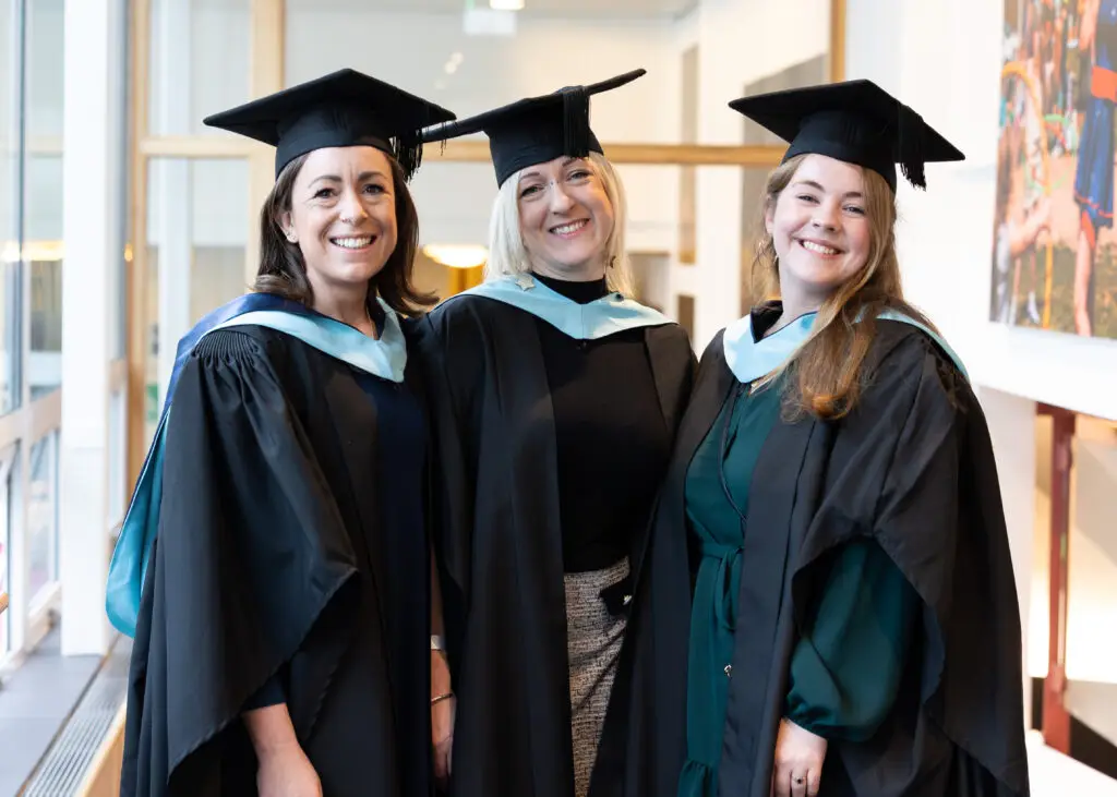 Three women in their graduation robes, standing side by side and smiling at the camera.