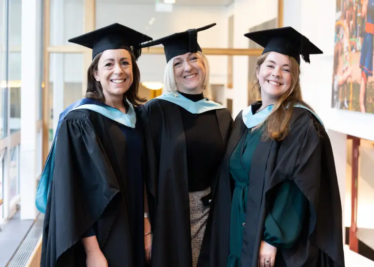 Three women in their graduation robes, standing side by side and smiling at the camera.