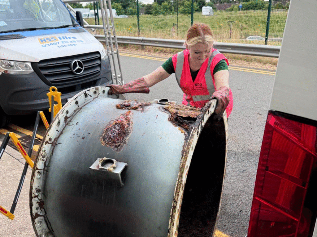 A woman wearing protective gloves and a high-visibility vest moves a large, rusted metal drum beside a work vehicle on an industrial site.