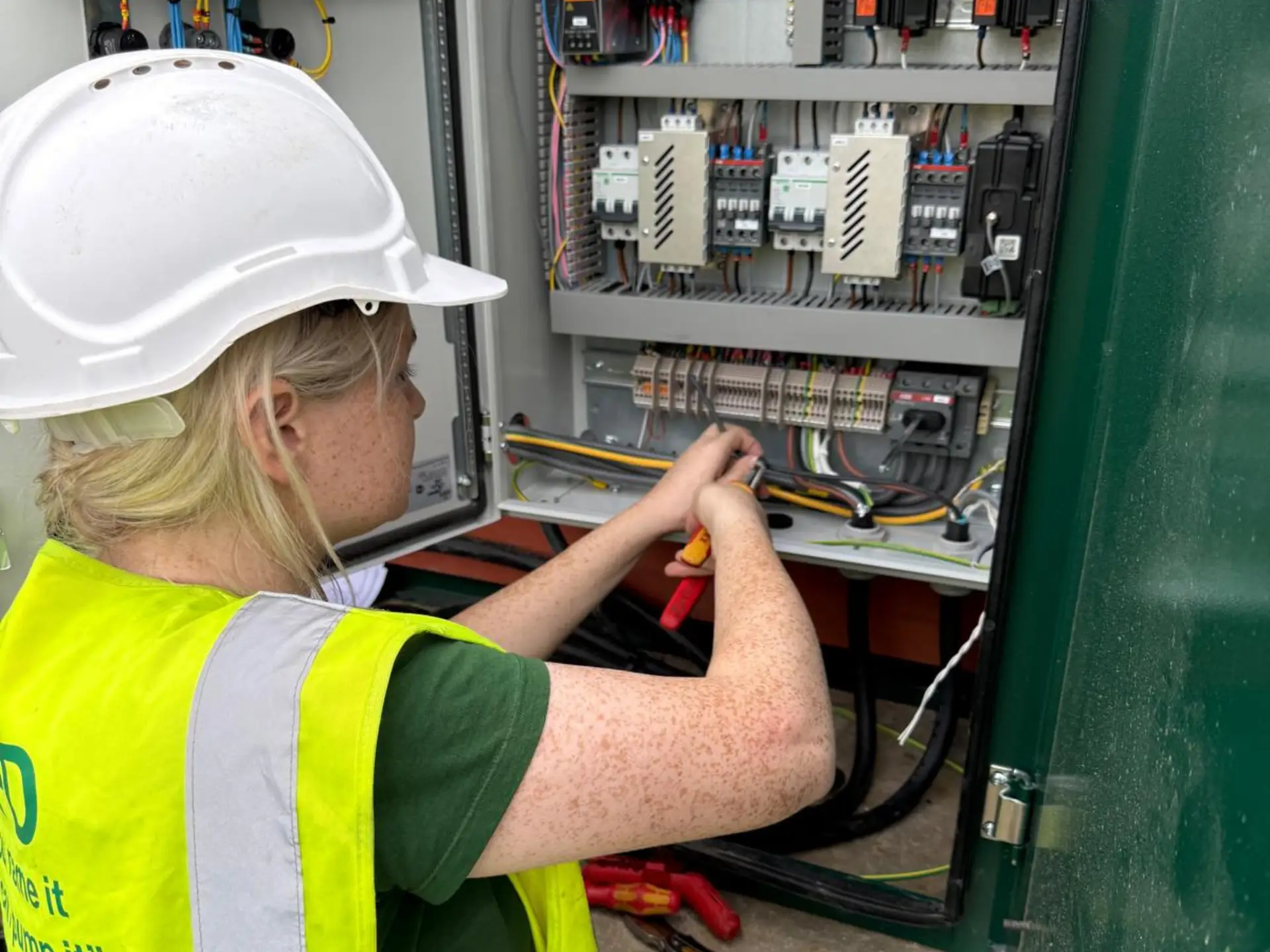 A woman wearing a hard hat and high-visibility vest uses tools to work on electrical wiring inside a large industrial control panel.