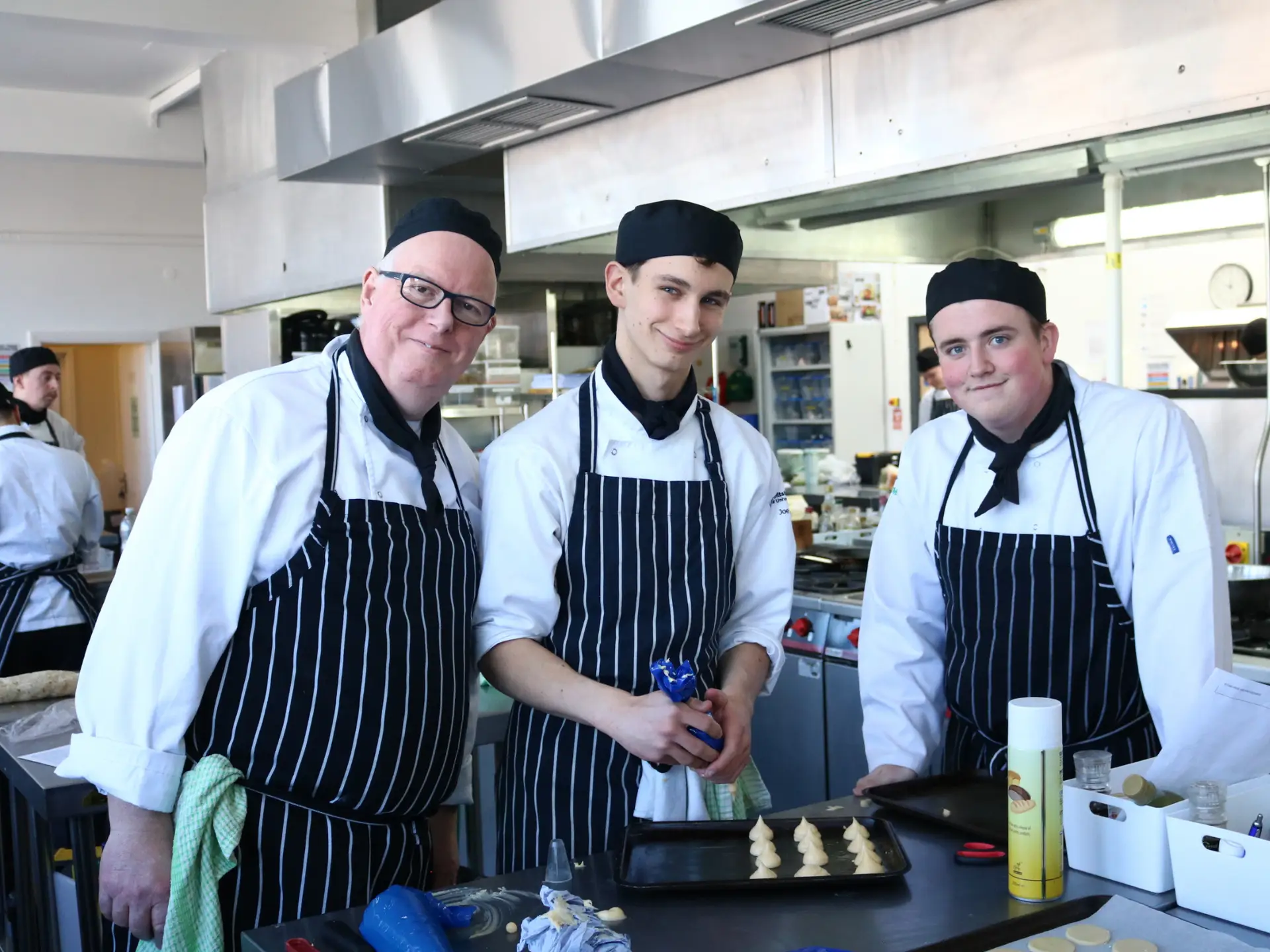 Hospitality students and a chef lecturer in a training kitchen preparing trays of piped pastry.