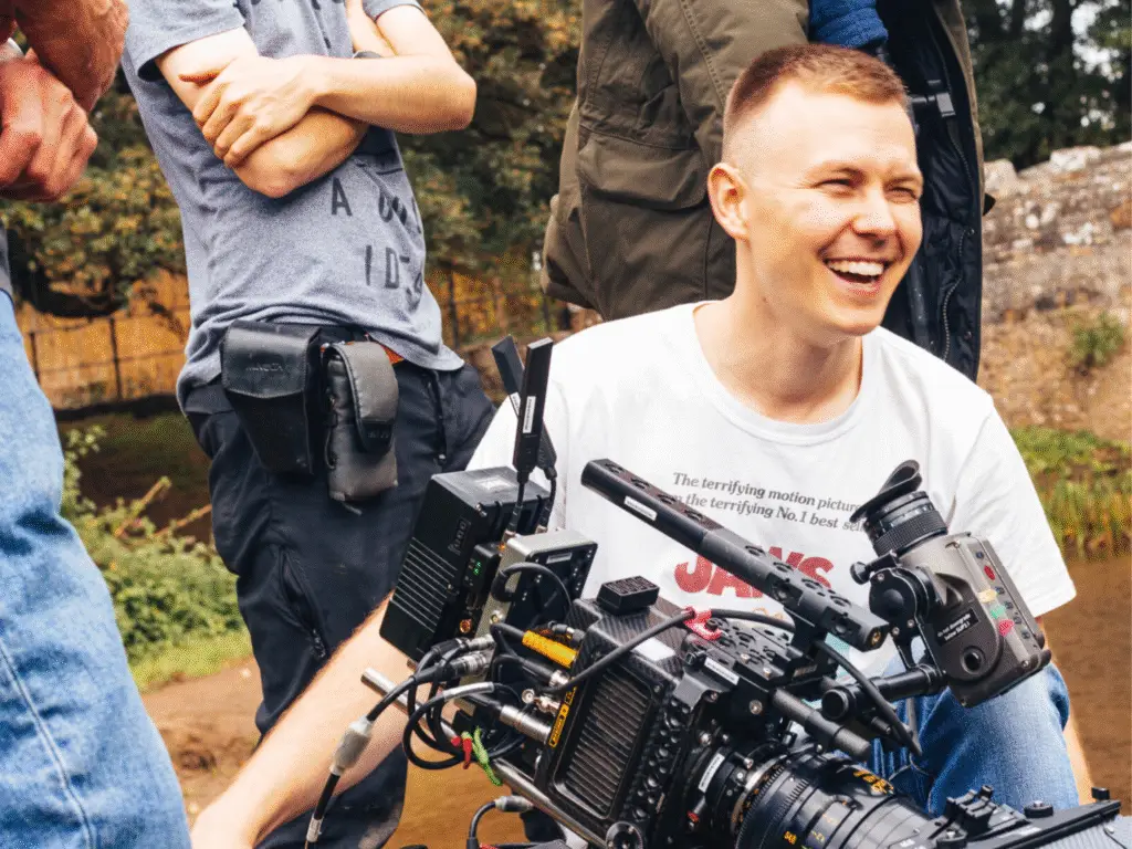 Student filmmaker laughing while operating a professional cinema camera during a film shoot outdoors.