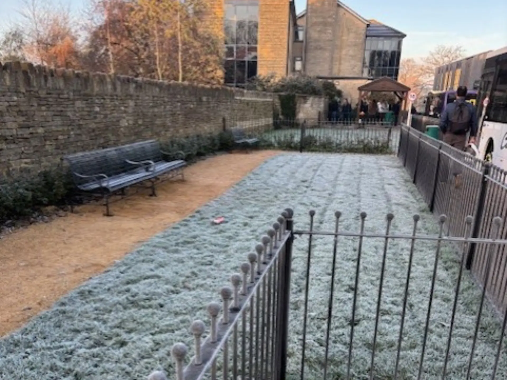 A frosty morning scene on campus showing a gravel path, metal benches and a grassed area covered in frost, with students waiting near buses in the background