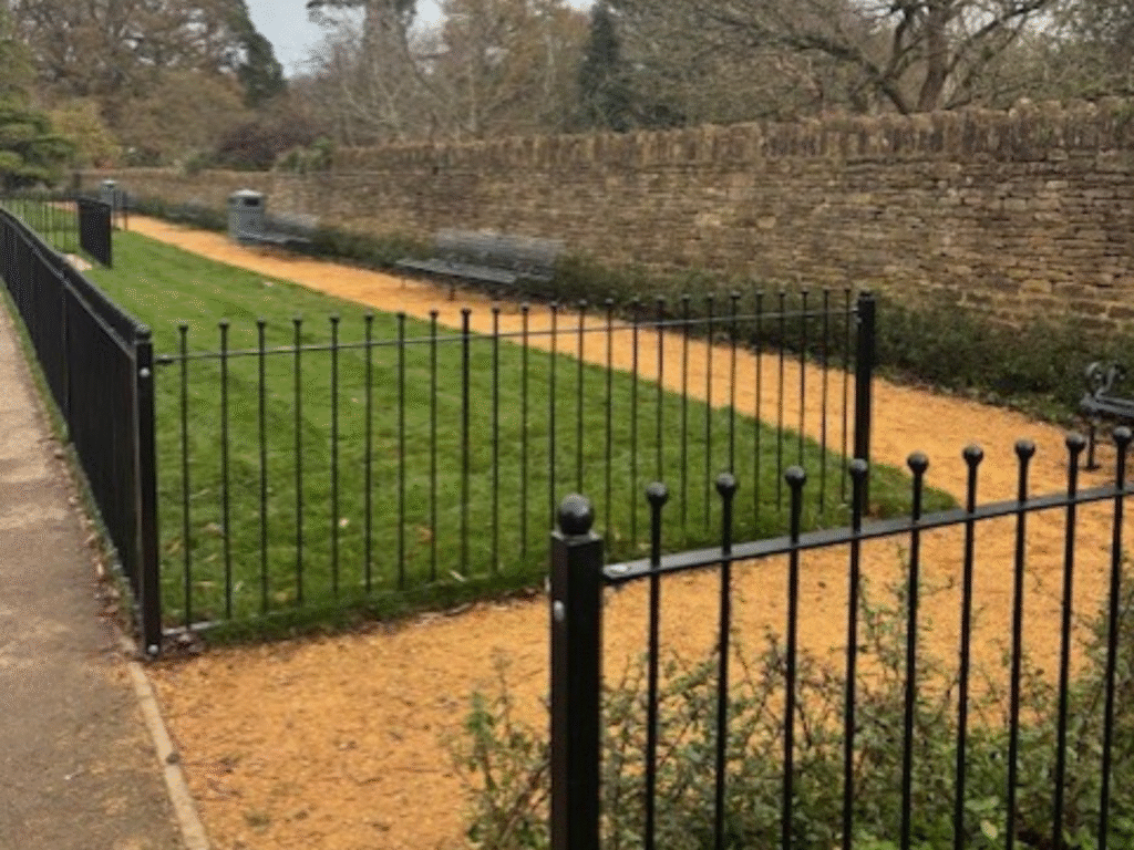 A landscaped campus area with newly laid grass, black metal fencing and a gravel pathway, bordered by a stone wall and benches.