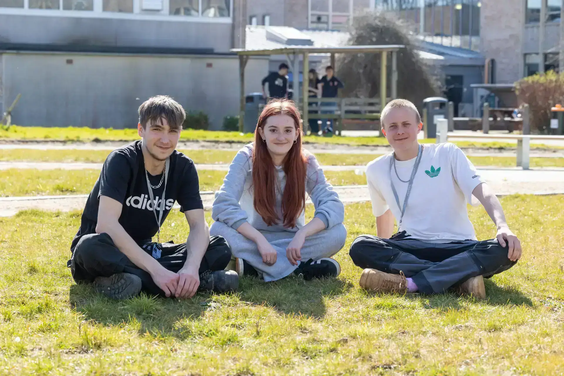 Three students sitting on the grass in the new courtyard at Wiltshire College & University Centre’s Trowbridge campus on a sunny day, with college buildings and other students visible in the background.