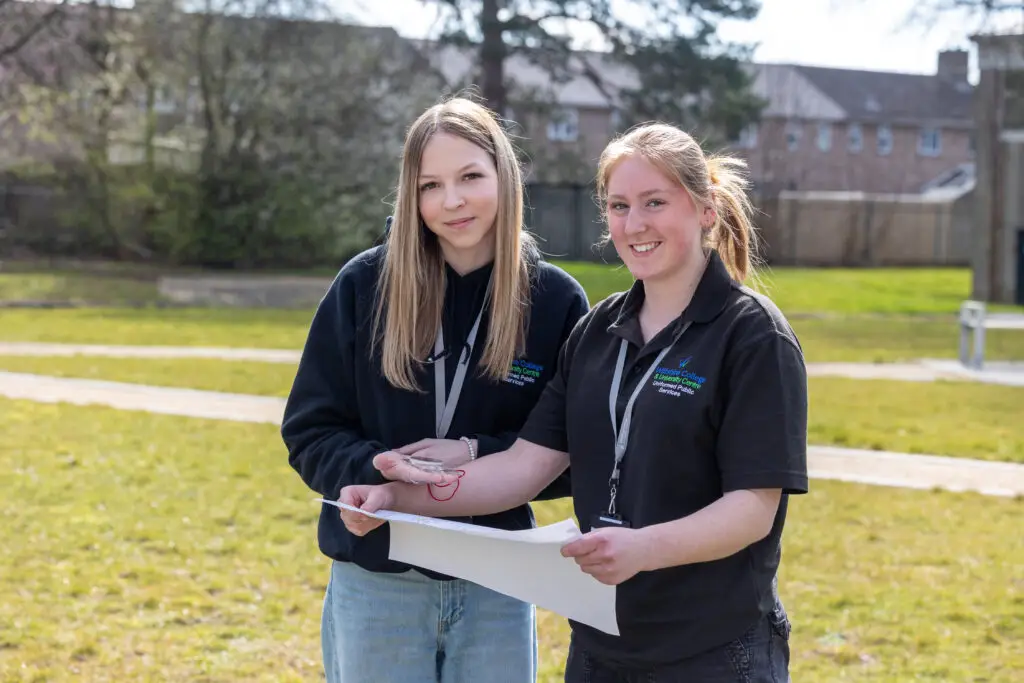Two students at Wiltshire College & University Centre’s Trowbridge campus standing in the new courtyard area, holding a map and smiling towards the camera on a sunny day.