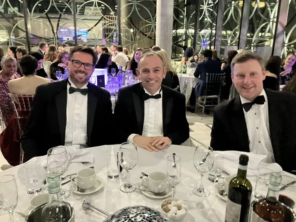 A group of three people dressed in formal evening wear seated at a round table during an awards ceremony.