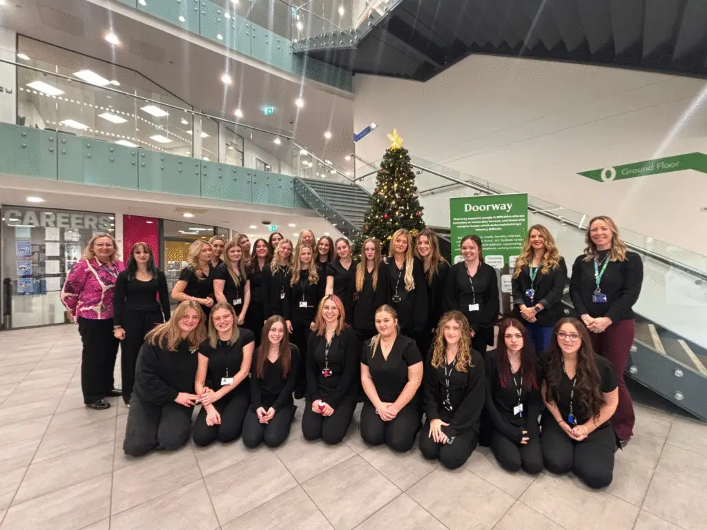 Hair and Beauty students and staff from Wiltshire College & University Centre standing together in the Chippenham campus atrium beside a Christmas tree and Doorway charity display.