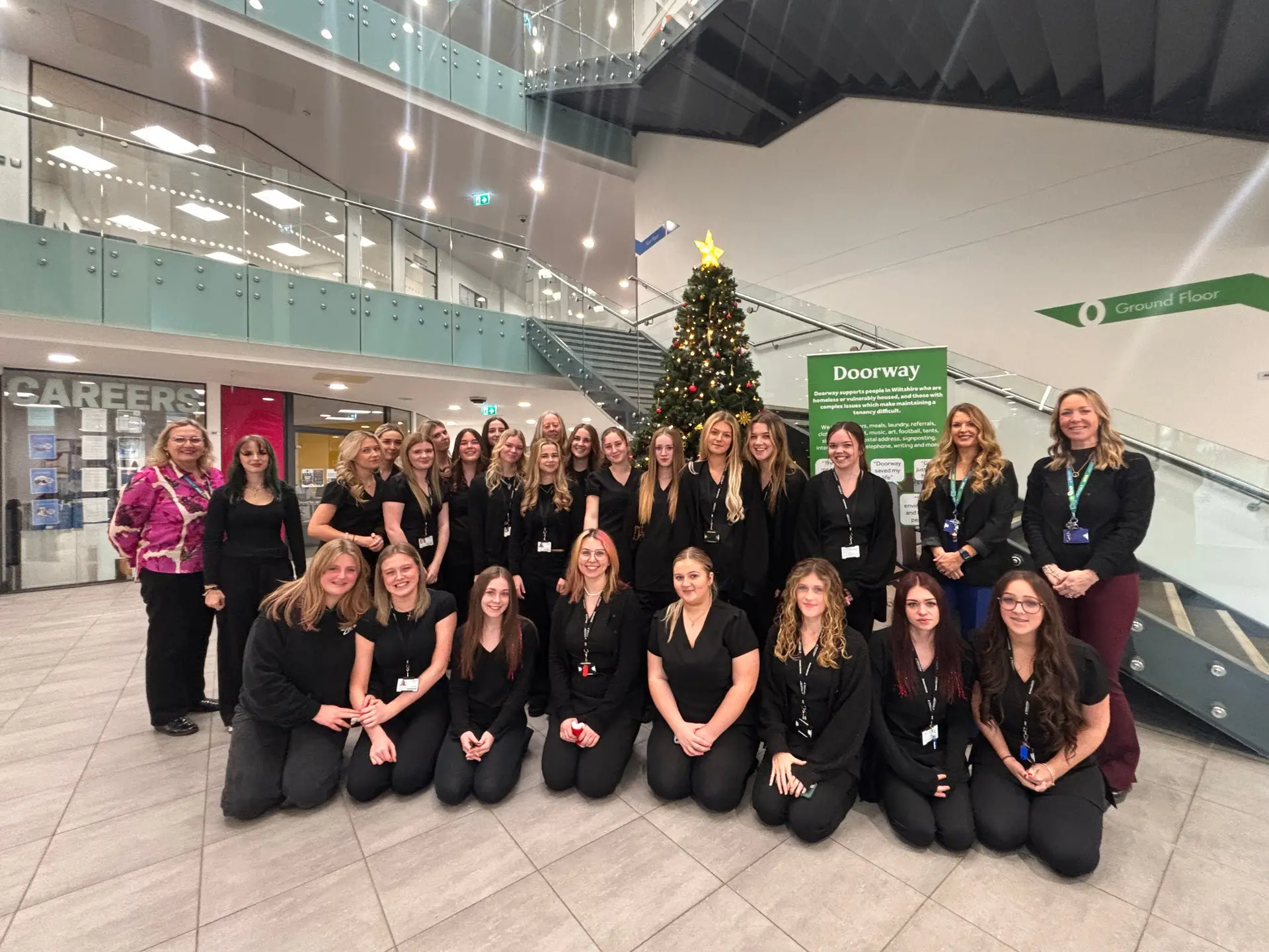 Hair and Beauty students and staff from Wiltshire College & University Centre standing together in the Chippenham campus atrium beside a Christmas tree and Doorway charity display.