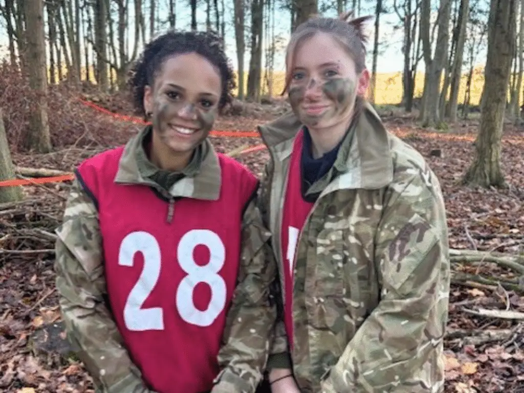 Two uniformed students wearing numbered training bibs stand together outdoors with camouflage face paint.
