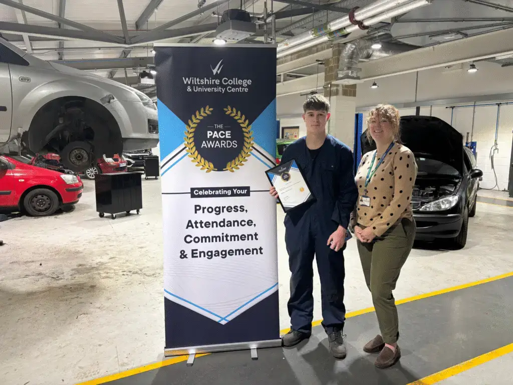 A student holding a PACE Award certificate stands beside a member of staff in an automotive workshop, with the Wiltshire College & University Centre PACE Awards banner displayed.