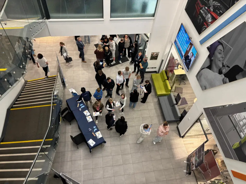 A busy open event in a college atrium with groups of visitors talking to staff near an information desk.