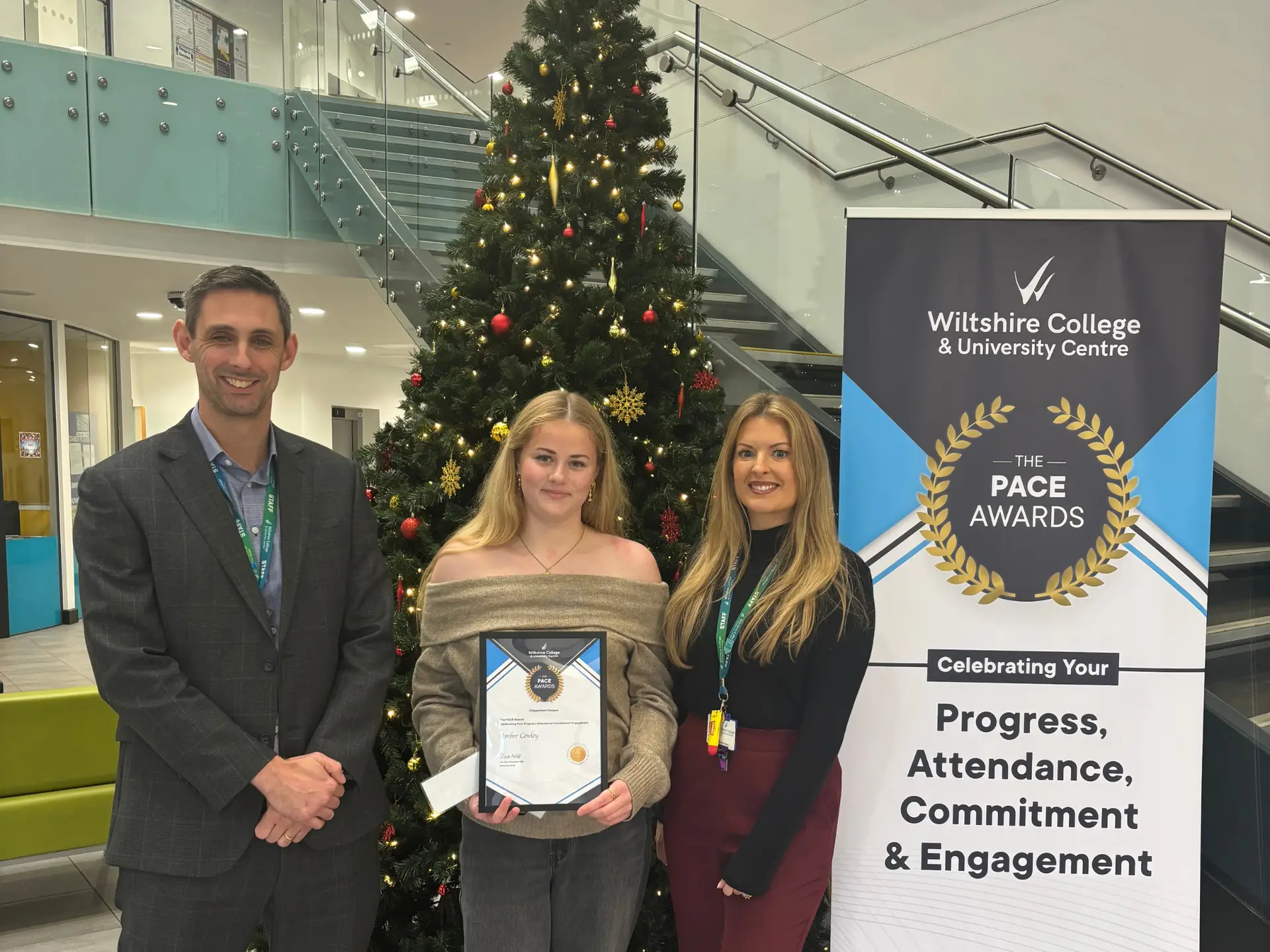 A student holding a PACE Award certificate stands between two members of staff beside a Christmas tree and a Wiltshire College & University Centre PACE Awards banner.