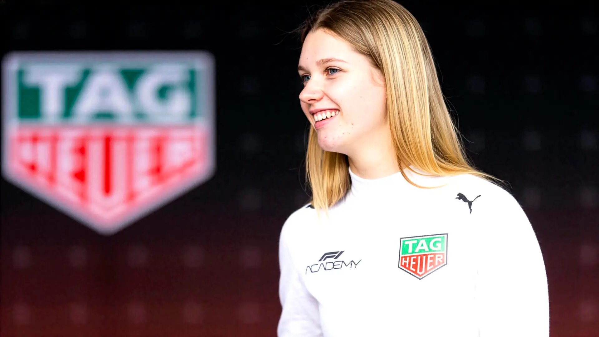 A young woman in a white motorsport team top smiling in front of a blurred sponsor backdrop.