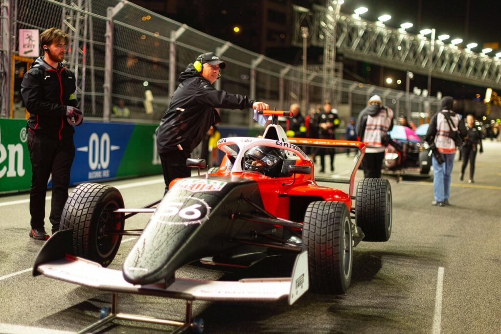 A single-seater race car sits on a lit street circuit at night with team members working around it.