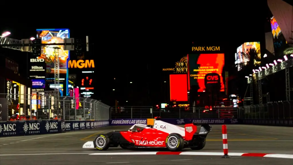 A red and black single-seater race car drives along a night-time street circuit with bright Las Vegas signage in the background.