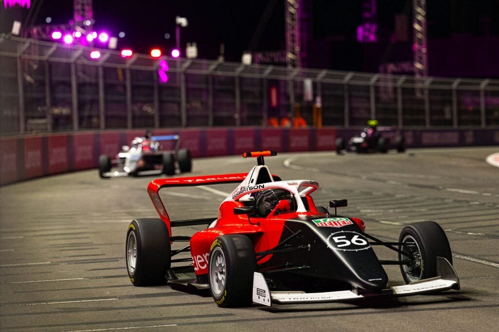 A red and black single-seater race car leads other cars through a corner on a night-time circuit.