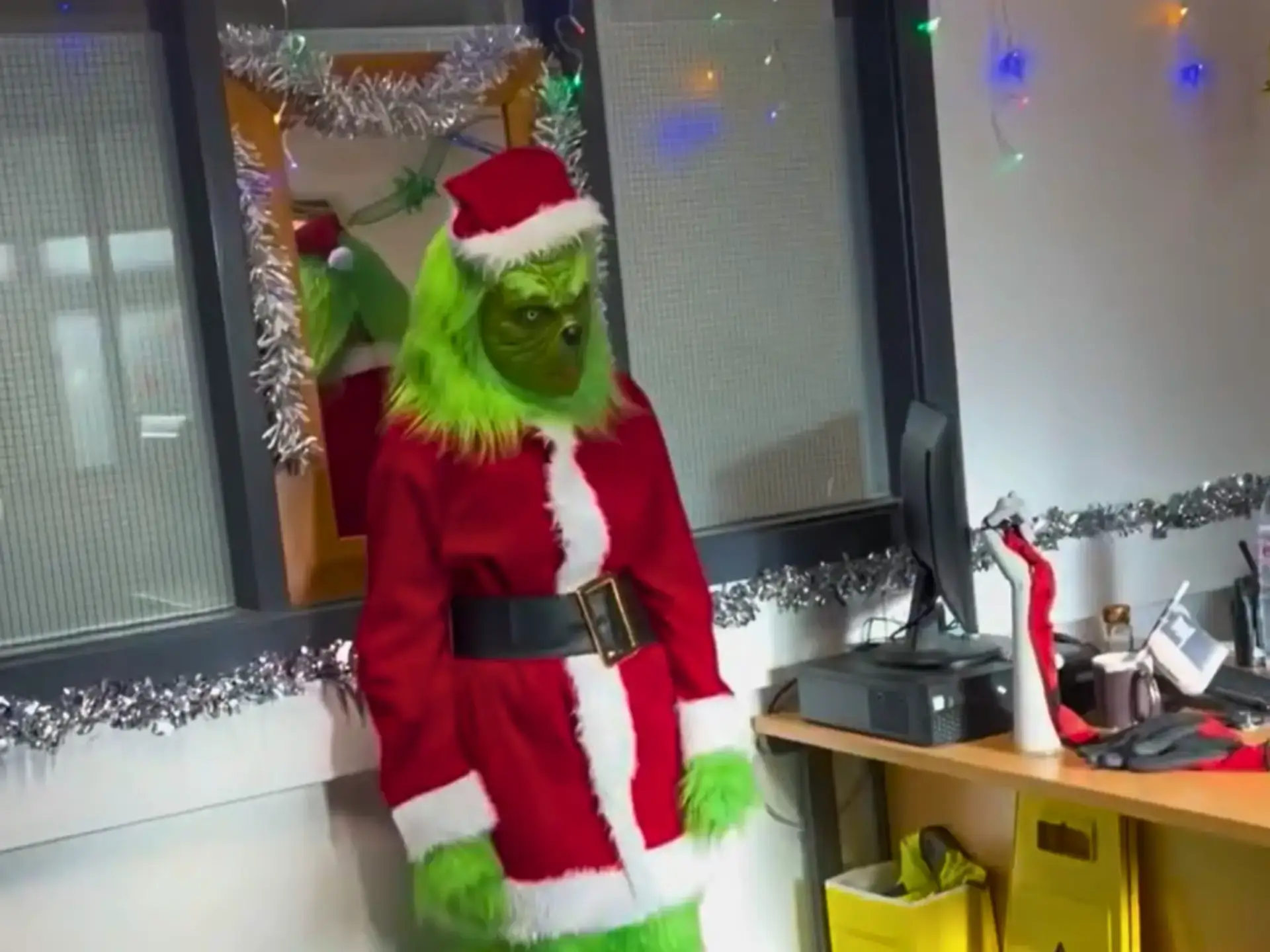 Student dressed as the Grinch wearing a Santa-style costume, standing in a festively decorated room on campus.
