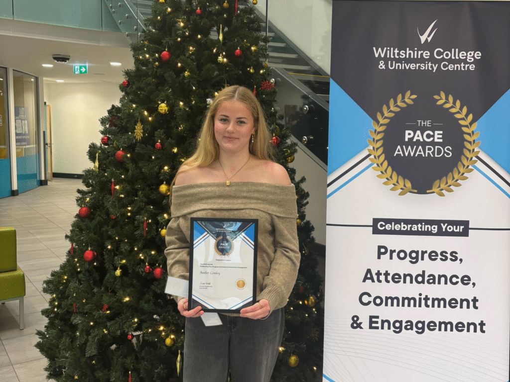 A student holding a PACE Award certificate stands in front of a Christmas tree beside a Wiltshire College & University Centre PACE Awards banner.